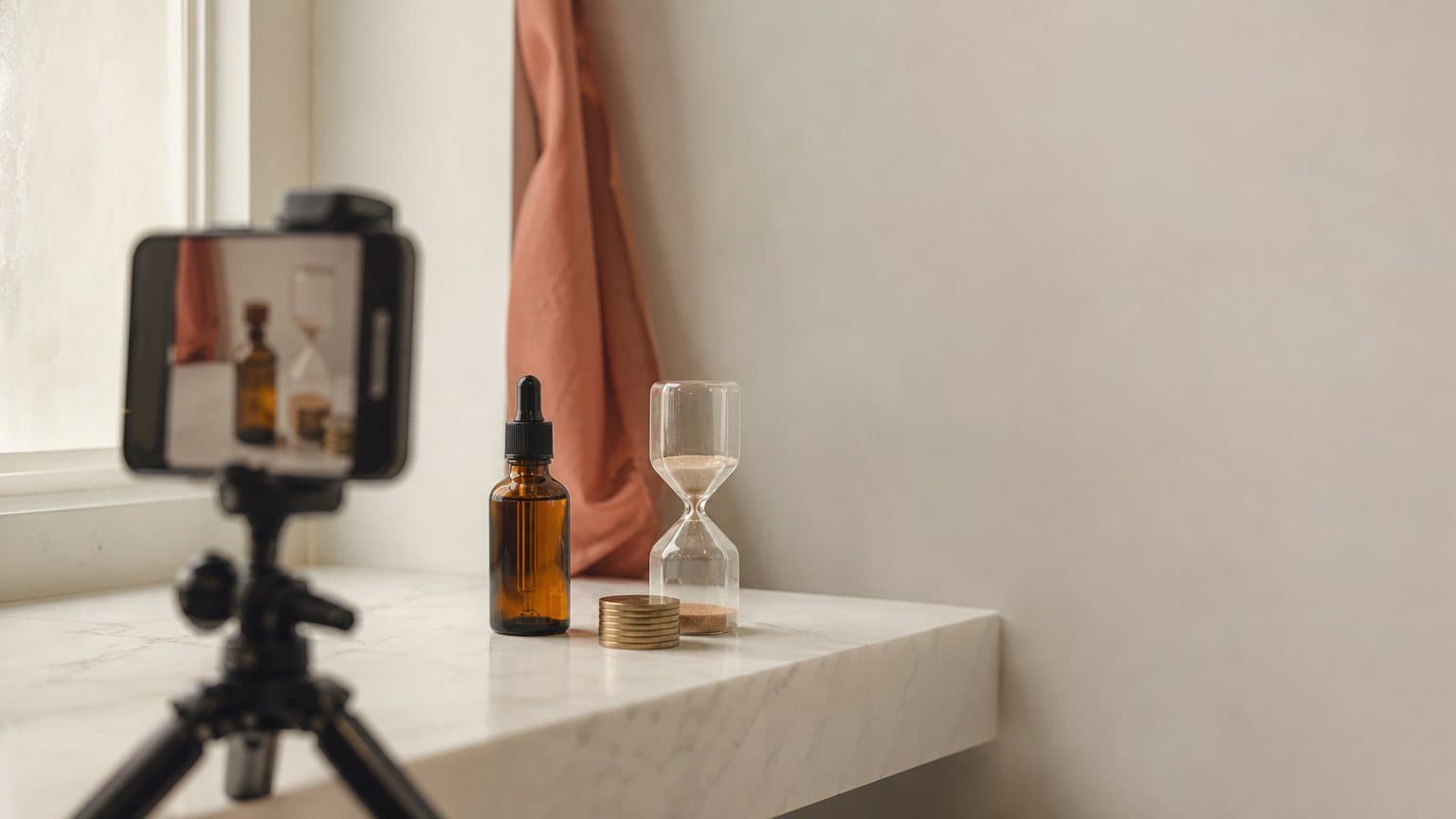 A smartphone on a small tripod filming an amber serum bottle, a glass hourglass and a short stack of brass tokens on a white marble bathroom shelf