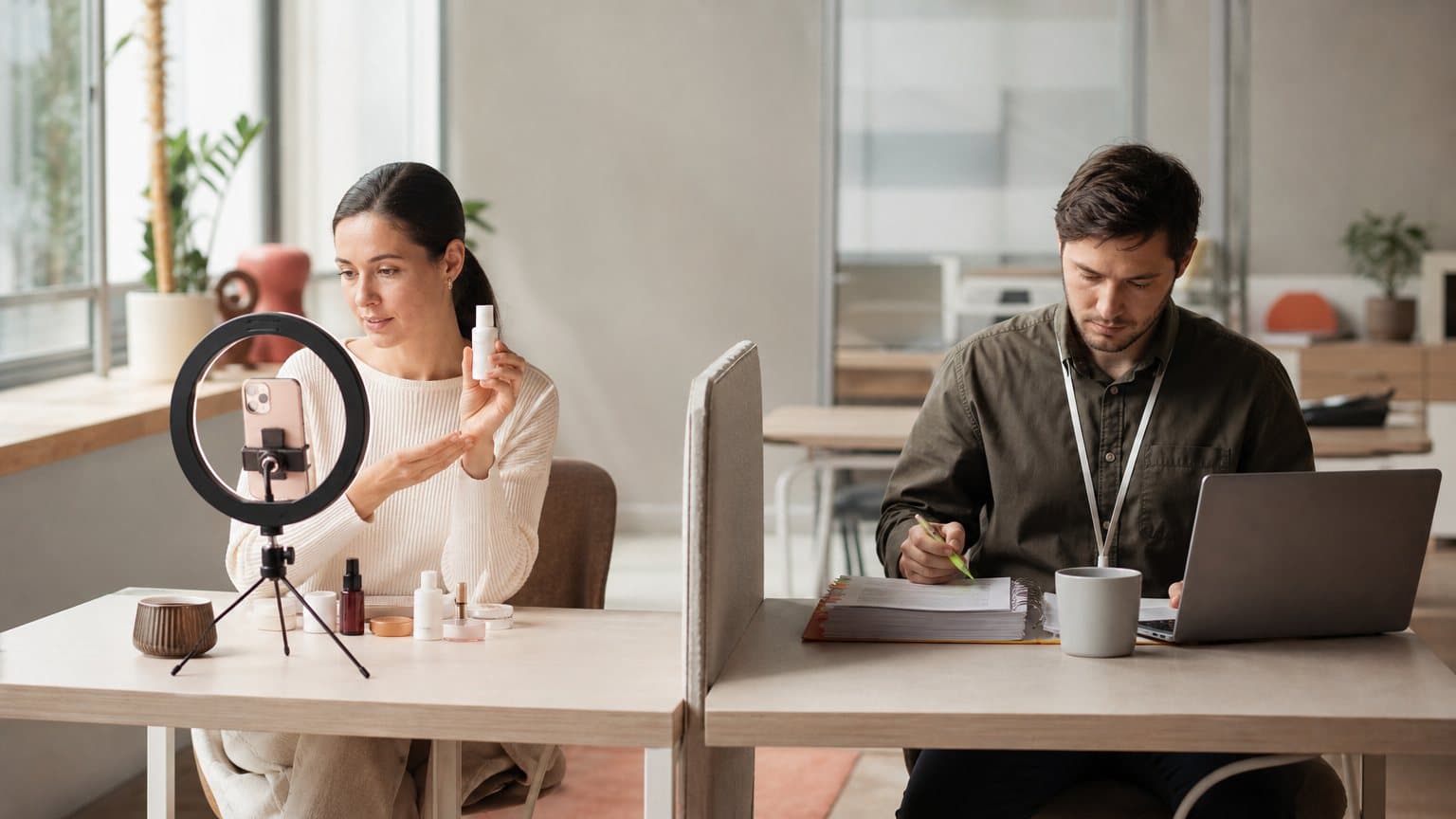 Two desks in one office: a marketer filming a bottle on a phone tripod beside a colleague working through an onboarding binder at a laptop